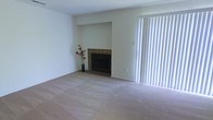 This single-floor space presents a clean, understated living area designed with simplicity and functionality in mind. The room is finished with neutral wall-to-wall carpeting in a warm beige tone, creating a soft and cohesive foundation throughout the space. A centerpiece fireplace, framed with a wood-trimmed surround and set into a recessed niche, serves as the room's focal point, adding a classic and cozy architectural detail to the otherwise minimal design. The white walls are kept clean and bright, contributing to an open and airy atmosphere. A large sliding door dressed with full-length vertical blinds occupies one wall, offering the ability to control natural light intake while suggesting access to an outdoor area. The vertical blinds diffuse incoming light softly across the room, enhancing the calm, neutral ambiance. A simple decorative vase with red floral stems near the fireplace adds a subtle touch of color and warmth to the otherwise unfurnished setting. The overall aesthetic is practical and move-in ready, offering a blank canvas well-suited for a variety of interior design preferences.