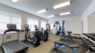 This fitness center offers a thoughtfully arranged workout space designed to support a full range of exercise routines. The room is finished with clean white walls and a suspended ceiling fitted with bright fluorescent lighting, ensuring excellent visibility throughout. Natural light also filters in through multiple windows dressed with white horizontal blinds, contributing to an open and energizing atmosphere.

The cardio section features multiple elliptical trainers positioned along the windowed wall, providing a comfortable area for sustained aerobic exercise. Rubber floor mats underneath the equipment offer stability and floor protection. A treadmill is also visible along the perimeter, rounding out the cardio offerings.

On the strength training side, a robust blue multi-functional cable machine serves as the centerpiece, offering a wide variety of resistance exercises. An adjustable weight bench in a matching blue frame is positioned nearby, accompanied by free weights, making it easy to transition between exercises.

The flooring throughout the space consists of patterned carpet tiles, adding a practical yet polished look underfoot. A doorway visible in the background suggests additional adjoining utility or storage space. Overall, the fitness center presents a clean, functional, and well-maintained environment suited for a variety of fitness goals.
