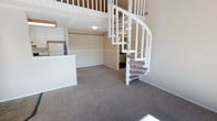 This two-floor residence offers a thoughtfully arranged layout that maximizes its vertical space through a standout white spiral staircase, which serves as both a functional connector between floors and a visual centerpiece of the main living area. The ground floor features an open-concept design where the living space flows naturally into the kitchen area. The living room is finished with wall-to-wall neutral-toned carpeting that lends a warm and comfortable feel underfoot. A breakfast bar or counter divides the kitchen from the living space, providing a casual dining or prep surface while maintaining the open sightlines of the main floor. The kitchen is equipped with standard appliances and cabinetry, offering practical storage and workspace. Neutral beige walls throughout the main level contribute to a clean, understated aesthetic. The upper level, accessible via the spiral staircase, overlooks the living area below, adding a sense of openness and height to the overall space. The two-story design creates a dynamic sense of volume, making the interior feel more expansive than a single-level layout might suggest. Overall, the space presents a functional and straightforward living environment with a distinctive architectural touch in the form of its spiral staircase.