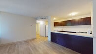 This single-floor residence at 10 Chestnut St in Springfield, Massachusetts offers a clean, modern interior defined by consistent light wood-tone flooring that flows seamlessly throughout the space. The open-concept layout connects the living area to the kitchen, which features dark espresso-toned cabinetry, light quartz-style countertops, a stainless steel sink, and white appliances including a gas range and refrigerator. A bold navy kitchen island base adds a distinctive design accent to the space. The living areas are generously sized, with large corner windows dressed in horizontal blinds that invite abundant natural light and offer views of the surrounding greenery. Neutral, light-toned walls contribute to a bright and airy ambiance throughout. The bathroom is well-appointed with white subway tile surrounding a full soaking tub and shower, a dark-cabinetry vanity with a white countertop, chrome fixtures, and a mirrored medicine cabinet. A second bathroom provides a toilet and vanity with matching finishes. The layout, as visible in the dollhouse view, includes distinct bedroom, living, kitchen, and bathroom zones arranged in a functional and accessible single-level floor plan, making this a well-organized and comfortable space.
