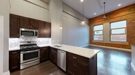 Located at 31 Barker St in Buffalo, NY, this two-story, two-bedroom, three-bathroom residence masterfully combines industrial character with contemporary design. The main floor makes an immediate impression with soaring ceilings that amplify the sense of openness throughout the living and kitchen areas. Exposed brick walls add warmth and texture, complemented by rich dark hardwood flooring and large windows that invite abundant natural light and street views. The kitchen is a standout, featuring dark espresso cabinetry, a mosaic tile backsplash, crisp white countertops, stainless steel appliances, and stylish pendant lighting that hangs dramatically from the tall ceiling. A peninsula provides additional prep and seating space. The upper floor houses the bedrooms, including a generously sized primary bedroom, all finished with plush carpeting. Closets offer practical storage, and the layout flows naturally to the staircase connecting both levels. The three bathrooms are finished in a clean, modern style — one featuring a full soaking tub and shower surround, another with a sleek floating vanity and tile flooring. Throughout, the combination of exposed brick, high ceilings, and thoughtful finishes creates a distinctive urban living environment.