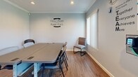 Located at 450 N Armando St in Anaheim, California, this single-room meeting space offers a clean and functional environment well-suited for small group gatherings, team meetings, or collaborative work sessions. The room is finished with warm wood-look laminate flooring and soft blue-gray walls accented by crisp white crown molding and baseboards, lending a polished yet approachable atmosphere throughout.

A central conference table with surrounding seating serves as the focal point of the space, accommodating several participants comfortably. A wall-mounted flat-screen television is positioned for easy viewing from the table, supporting presentations and media playback. A white media console below the TV provides additional storage and equipment housing.

Natural light filters in through windows fitted with vertical blinds, while recessed ceiling lights ensure consistent illumination. The room also features a wall-mounted HVAC unit for climate control. A glass-paned exterior door opens to the outside, adding to the sense of connectivity with the surrounding environment.

Decorative touches include a framed black-and-white coastal photograph and motivational wall decals, contributing to an energizing yet professional tone. A classic red popcorn machine adds a casual, welcoming element to the corner of the room. Security features including a fire extinguisher and access control device are also present, reflecting thoughtful preparation for a working environment.