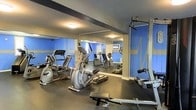 This fitness room is designed to support a well-rounded workout routine within a efficiently organized space. The room features bold blue walls adorned with graphic heartbeat-style accents, giving the space an energetic and motivating atmosphere. A large wall mirror spans a significant portion of the room, visually expanding the space while allowing users to monitor their form during exercise. The flooring consists of dark rubber matting, providing cushioning and durability suited to high-traffic workout use. Cardio equipment includes an elliptical trainer and a recumbent stationary bike, both positioned to make practical use of the available floor space. A multi-function cable machine is situated along one wall, offering options for strength and resistance training, while a flat workout bench adds further versatility. A wall-mounted television is visible in the mirror's reflection, providing entertainment during workouts. Overhead flush-mount lighting ensures the room is well-lit throughout. The overall ambiance is functional and purpose-driven, catering to residents or guests seeking convenient access to fitness amenities.