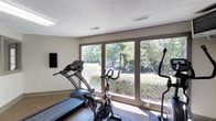 This single-floor fitness space is thoughtfully designed to support an active lifestyle in a bright, welcoming environment. The room is anchored by a bank of large picture windows that span nearly the full width of one wall, providing sweeping views of mature trees, manicured shrubbery, and a lush green landscape beyond. The abundance of natural light that pours through these windows creates an airy, open atmosphere that makes workouts more enjoyable.

The fitness area is equipped with essential cardio equipment, including a treadmill, a stationary spin bike, and an additional exercise machine, offering a well-rounded selection for cardiovascular training. A wall-mounted display panel adds a modern, functional touch to the space. The flooring features a durable, textured surface suited for exercise use, providing stability and comfort underfoot.

The ceiling features a textured finish with a centrally positioned ventilation duct, ensuring proper airflow throughout the room. Neutral-toned walls keep the environment clean and focused, allowing the natural scenery outside to serve as the visual centerpiece. Overall, this fitness room combines practical amenities with a bright, nature-connected setting to deliver an energizing and motivating workout experience.