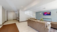 Located at 556 Colonial Rd in Franklin Lakes, New Jersey, this residential building lobby serves as an inviting and well-appointed entryway for its residents. The single-floor hallway space is thoughtfully designed to balance functionality with comfort.

Upon entry through the large glass front doors, natural light floods the space, illuminating the clean, large-format cream tile flooring accented by a warm brown runner that guides visitors inward. The entry zone features wall-mounted sconces and recessed ceiling lighting throughout, creating a bright and consistent ambiance at all hours.

The lounge area is a standout feature, furnished with a set of tan leather sofas and armchairs arranged around a modern wood-and-metal coffee table atop a colorful geometric area rug. A wall-mounted flat-screen TV is set against a stylish textured blue-gray accent wall, adding a contemporary touch to the space.

Adjacent to the lounge, a dedicated mailbox station provides organized mail access for all residents, accompanied by a console table with a decorative lamp. Artwork adorns the walls, contributing to the polished, welcoming atmosphere. An elevator and interior corridor door provide seamless access to the rest of the building. Large windows along one wall allow additional natural light and views of the surrounding greenery, enhancing the overall sense of openness.
