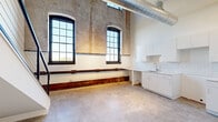 Located at 356 Hertel Ave in Buffalo, NY, this two-bedroom, two-bathroom loft blends industrial heritage with modern finishes across two floors. The main level features polished concrete floors, soaring ceilings with exposed ductwork, and original brick and concrete walls that preserve the building's historic character. Large arched and grid-pane windows flood the space with natural light while framing views of the surroundings. The open-concept kitchen, dining, and living area is anchored by white shaker cabinetry, a subway tile backsplash, and a continuous countertop that wraps the kitchen. A sleek staircase with black metal railings leads to the upper level, where warm hardwood floors complement the more intimate atmosphere. The spacious primary bedroom is accessed through a signature frosted-glass sliding barn door with a dark metal frame — a design detail echoed throughout the home. Both bathrooms are finished in crisp white, with the primary bath featuring marble-look tile floors, subway tile walls, a frameless glass shower, and a white vanity. A dedicated laundry area and hallway complete the layout. The result is a thoughtfully converted loft that honors its industrial roots while delivering clean, contemporary livability.