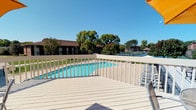 This single-floor facility is designed to serve as a comprehensive recreational destination, offering both outdoor amenities and functional interior spaces. The outdoor area is a standout feature, centered around a generously sized swimming pool with crystal-clear blue water, surrounded by a wide, light-colored composite deck finished with a white balustrade railing. The deck provides open views of the lush, tree-lined surroundings and connects seamlessly to tennis courts visible in the background, making this an ideal multi-sport venue. A partial shade umbrella adds a practical touch to the poolside experience, and the overall outdoor environment benefits from abundant natural light under open skies. The interior restroom facility is thoughtfully designed for high-traffic use, featuring multiple side-by-side stall units equipped with stainless steel fixtures, sensor-activated faucets, and warm wood-toned cabinetry beneath each basin. The restroom stalls are fitted with automated flush mechanisms and neatly organized lower shelving, reflecting a clean and utilitarian design. The interior finishes maintain a neutral palette, contributing to a clean and functional ambiance throughout. Together, the outdoor and indoor spaces form a cohesive recreational environment well-suited for community or resort use.