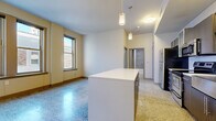 Located at 306 E 12th St in Kansas City, MO, this two-bedroom, two-bathroom unit offers a well-designed single-floor layout with a clean, modern aesthetic throughout. The open-concept kitchen serves as a central hub, featuring dark cabinetry with glass-front upper cabinets, a white quartz countertop island with pendant lighting, a subway tile backsplash, and a full suite of stainless steel appliances. Polished terrazzo flooring extends through the main living and kitchen areas, adding a distinctive, durable finish. The primary bedroom is generously sized with plush carpeting, large windows showcasing urban Kansas City views, and a dedicated walk-in closet with wire shelving. The second bedroom also features carpet, a built-in closet, and bright, neutral walls. Both bathrooms are well-appointed: one features a bold teal accent wall, a floating white vanity, and a tiled tub-surround, while the other offers a clean, tiled shower-tub combo with coordinating grey tile. A hallway with additional closet access connects the private quarters. The unit benefits from elevated ceilings, ample natural light from city-facing windows, and a cohesive contemporary design throughout.