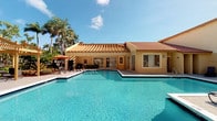 This single-story property showcases a Mediterranean-inspired architectural style, most notably highlighted by its distinctive terracotta tile roof and warm yellow stucco exterior. The standout feature of the outdoor space is a generously sized swimming pool with vivid turquoise water, which serves as the centerpiece of the property's exterior. The pool area is surrounded by a wide, clean patio deck that provides ample space for outdoor seating and leisure activities. A patio dining set with an orange umbrella is positioned poolside, offering a shaded area for relaxation. Large sliding glass doors and windows line the rear-facing exterior wall of the building, facilitating a strong visual and physical connection between the interior and the outdoor pool area, while also allowing abundant natural light into the interior spaces. Tall, mature palm trees frame the property, contributing to a lush, tropical ambiance that complements the overall Mediterranean aesthetic. The combination of the warm exterior tones, vibrant pool, and tropical greenery creates an inviting, resort-like atmosphere. Note that interior images were not available for review, so details regarding interior rooms and finishes are limited to what is visible through the exterior windows.