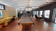 This single-floor amenity lounge is designed to accommodate a variety of social and recreational activities within one open, well-appointed space. The layout is anchored by a full-size billiards table with ornate carved legs, positioned beneath a statement barrel-style chandelier that adds a warm, industrial-rustic character to the room. Exposed beam detailing on the white ceiling complements the polished concrete flooring, which carries a rich, warm tone throughout the space.

Structural columns punctuate the open floor plan, subtly defining distinct zones without enclosing them. A comfortable lounge area with upholstered seating and a wall-mounted flat-screen television is positioned toward the rear, creating an inviting spot for relaxed gatherings. A shuffleboard table is visible along one side wall, adding another layer of entertainment options.

To one side, a large dining table surrounded by chairs provides space for group meals or casual meetings. Natural light filters in through multiple windows along the perimeter, contributing to the bright and airy atmosphere. Exposed brick accents on one wall add texture and a loft-like aesthetic, reinforcing the space's modern-industrial design sensibility.