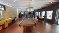 This single-floor amenity lounge is designed to accommodate a variety of social and recreational activities within one open, well-appointed space. The layout is anchored by a full-size billiards table with ornate carved legs, positioned beneath a statement barrel-style chandelier that adds a warm, industrial-rustic character to the room. Exposed beam detailing on the white ceiling complements the polished concrete flooring, which carries a rich, warm tone throughout the space.

Structural columns punctuate the open floor plan, subtly defining distinct zones without enclosing them. A comfortable lounge area with upholstered seating and a wall-mounted flat-screen television is positioned toward the rear, creating an inviting spot for relaxed gatherings. A shuffleboard table is visible along one side wall, adding another layer of entertainment options.

To one side, a large dining table surrounded by chairs provides space for group meals or casual meetings. Natural light filters in through multiple windows along the perimeter, contributing to the bright and airy atmosphere. Exposed brick accents on one wall add texture and a loft-like aesthetic, reinforcing the space's modern-industrial design sensibility.
