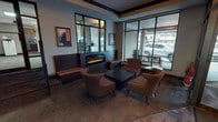 This single-floor lobby serves as the welcoming entrance to a residential building, combining functional design with warm, inviting aesthetics. Upon entry, visitors are greeted by a bold terracotta-orange accent wall adorned with abstract artwork, setting a contemporary tone. The glass-panel entry doors are framed with signage, offering an immediate sense of identity and place.

The lounge area is a standout feature, centered around a sleek, linear electric fireplace set within a dark wood surround and flanked by built-in bench seating with warm-toned cushions. Surrounding a low square coffee table, a cluster of curved, upholstered armchairs in rich brown leather invites relaxed conversation or quiet rest. Framed artwork decorates the walls throughout, adding a curated, gallery-like quality to the space.

Large floor-to-ceiling windows and glass walls flood the lounge with natural light while offering open views of the surrounding streetscape. The flooring throughout features a rich, patterned wood-look tile that adds texture and warmth underfoot.

The main hall connects to multiple staircase landings with classic iron railings, providing access to other areas of the building. Decorative ceiling fixtures cast a soft ambient glow, complementing the structured crown molding that runs throughout, tying the overall design together with a polished, cohesive finish.