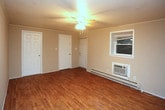 This single-floor 1-bedroom unit offers a practical and comfortable living space designed with functionality in mind. The main living area features warm hardwood-style flooring that extends throughout, providing a clean and cohesive look underfoot. A ceiling fan with an integrated light fixture is centrally mounted on the textured ceiling, offering both air circulation and ambient lighting. Climate control is well-covered with a baseboard heating system running along the wall and a wall-mounted air conditioning unit positioned beneath the window, ensuring comfort across all seasons. The window allows natural light to filter into the space, offering a view of the exterior brick surroundings. Multiple interior doors provide access to additional rooms and storage areas, including what appears to be a closet with double doors — a practical addition for everyday organization. The walls are finished in a neutral, light tone that keeps the space feeling open and airy. Overall, this unit presents a straightforward, well-maintained living environment with essential amenities thoughtfully in place.