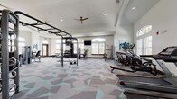 This single-story fitness facility is thoughtfully designed to support a wide range of workout routines. The main gym area is expansive and bright, featuring vaulted ceilings, multiple ceiling fans, and abundant natural light streaming through arched windows along the perimeter walls. The floor is covered in a bold geometric-patterned carpet in shades of gray and aqua, adding a modern, energetic aesthetic to the space. Equipment includes TRX functional training rigs, a heavy punching bag, pull-up stations, cable machines, weight benches, a full dumbbell rack, and an array of cardio machines such as treadmills, ellipticals, and stationary bikes. A wall-mounted flat-screen television is positioned for easy viewing throughout the gym floor.

Adjacent to the main gym is a dedicated yoga and movement studio, featuring warm hardwood flooring, high ceilings with decorative tray detailing, framed abstract artwork on clean white walls, and ample space for group or individual practice. Exercise mats and stacked props are available within the room. The entry foyer connecting the spaces is finished with large-format tile flooring, soft teal-painted walls, a cushioned bench, and a shelving unit stocked with rolled yoga mats, providing an organized and welcoming transition into the facility.