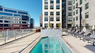 Located at 3720 N Downing Street in Denver's vibrant Cole neighborhood, this rooftop pool and spa amenity offers a well-appointed outdoor retreat with striking urban views. The centerpiece is a generalist lap pool finished with clean, light-toned tile and clear blue water, complemented by a separate spa encased in a distinctive mosaic tile surround wall. An accessible pool lift is available, underscoring the space's inclusive design. Surrounding the water features, a generous sun deck is furnished with multiple rows of modern slatted chaise lounges, providing ample space for relaxation. A pergola with a slatted wood ceiling offers a shaded gathering area adjacent to the pool, adding architectural warmth to the otherwise contemporary setting. The deck is enclosed by a combination of black iron fencing and glass panel railings, maintaining safety while preserving unobstructed sightlines. From nearly every vantage point, the Denver city skyline and the Rocky Mountain backdrop are visible on the horizon, making this outdoor space as visually compelling as it is functional. The elevated setting bathes the entire area in natural sunlight, creating a bright and open atmosphere ideal for outdoor leisure in the heart of Denver.