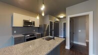 Located at 115 E 5th Ave in Columbus, Ohio's dynamic Short North neighborhood, this single-floor unit offers a thoughtfully designed modern living space with high-quality finishes throughout. The kitchen is a standout feature, showcasing granite countertops, gray cabinetry, a large island with an undermount sink, and a full suite of stainless steel appliances including a refrigerator, range, microwave, and dishwasher. Edison bulb pendant lighting and a geometric tile backsplash add a contemporary industrial touch. Dark hardwood-style flooring flows seamlessly through the kitchen and into the adjoining living area, which is bathed in natural light and opens directly onto a private balcony through a sliding glass door, offering outdoor views of the surrounding neighborhood. The bedroom is a calm, comfortable retreat featuring a ceiling fan, a large window with natural light, and the same warm hardwood-style flooring. The bathroom is well-appointed with a granite-topped vanity, dark cabinetry, and a glass-enclosed walk-in shower with elegant marble-style tile. An in-unit stacked washer and dryer provide added convenience. The space is finished with neutral gray tones, clean white trim, and modern fixtures throughout, creating a cohesive and polished aesthetic.