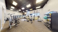 This fitness center offers a spacious, single-floor layout designed to accommodate a comprehensive range of workout needs. The room features impressively high ceilings adorned with a distinctive dark grid-patterned tile ceiling with recessed panel lighting, creating a bold and energizing atmosphere. The flooring consists of durable, neutral-toned carpet tiles throughout, providing comfort and stability during exercise.

Natural light fills the space through multiple large windows, including a prominent arched window, complemented by roller shades for adjustable light control. Views of palm trees and the surrounding outdoor environment add a refreshing ambiance to the workout experience. Wall-mounted flat-screen televisions are positioned throughout for entertainment during cardio sessions.

The cardio zone is well-appointed with treadmills, elliptical trainers, stationary bikes, and a StairMaster rowing machine. The strength training area includes a multi-station cable machine, a power tower, a full dumbbell rack, weight benches, and a dedicated medicine ball and stability ball station. Full-length mirrors are strategically placed to support proper form and technique.

The space also features a separate ancillary corridor visible near the entrance, suggesting additional support facilities. Overall, the fitness center presents a clean, professional environment well-suited for both cardio and strength training routines.