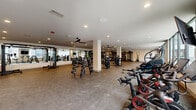 This expansive, single-floor fitness center offers a comprehensive workout environment designed to accommodate a wide range of exercise disciplines. The space is thoughtfully organized into distinct functional zones, including a cardio area, a free weights section, and a strength training zone, allowing users to move seamlessly between equipment.

The cardio area features a row of treadmills and elliptical machines positioned alongside large floor-to-ceiling windows that flood the space with natural light and provide views of the surrounding exterior. Ceiling fans are installed throughout, ensuring proper air circulation across the facility. The strength training zone is generously outfitted with cable machines, multi-functional rack systems, and wall-mounted mirrors that visually expand the space and aid in form monitoring.

The free weights section includes a full rack of dumbbells, barbells, kettlebells, medicine balls, and stability equipment, catering to both beginner and advanced training routines. Peloton bikes and a RowERG rowing machine round out the cardio offerings for low-impact, high-intensity options. The flooring throughout consists of durable rubber and carpet surfaces suited for heavy equipment and high-traffic use.

Recessed lighting combined with the abundant natural light creates a bright, energizing atmosphere. The clean, neutral color palette and modern finishes contribute to a professional and welcoming environment ideal for daily fitness routines.