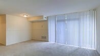 This single-floor space features a open living area finished with wall-to-wall carpeting in a neutral tone, contributing to a clean and understated aesthetic. The room is characterized by its large sliding glass doors, which span nearly the full width of one wall and are fitted with white vertical blinds. These doors allow an abundance of natural light to filter into the space while also providing direct outdoor access. The walls are painted in a soft, light cream tone, complementing the bright and airy atmosphere. A stepped ceiling detail adds subtle architectural interest, creating a partial soffit that defines a portion of the room. Practical features include a wall-mounted HVAC vent near the baseboard, a ceiling-mounted light fixture, and a visible thermostat or switch plate on the wall. The overall layout feels open and functional, with a simple, neutral palette that offers versatility for a variety of furnishing styles. The combination of natural light, access to the outdoors, and straightforward design makes this a practical and adaptable living space.