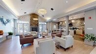 This well-appointed single-floor community space blends contemporary design with warm, inviting elements to create a sophisticated gathering environment. The centerpiece of the main lounge is a soaring stacked-stone fireplace that rises to a vaulted ceiling, flanked by large French doors that flood the space with natural light and provide views to the outdoors. Circular pendant light fixtures hang overhead, complementing the light hardwood flooring and neutral color palette of soft grays and whites.

The lounge area offers generous seating arrangements with a mix of plush sofas and accent chairs in cream and warm brown tones, ideal for both relaxed socializing and casual meetings. A striking blue sculptural wall installation adds an artistic focal point, lending the space a curated, gallery-like quality.

Adjacent to the lounge, a kitchenette area features a reclaimed wood accent wall, white countertops, stainless steel appliances including a microwave, refrigerator, and coffee station, all mounted beneath wall-mounted flat-screen televisions. A separate workspace nook provides high-top bar seating with ample counter space and natural light from large windows. A glass-walled conference or private meeting room offers additional functionality, completing this versatile and thoughtfully designed communal space.