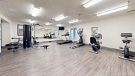 Located at 200 Valencia Dr SE in Albuquerque, New Mexico, this fitness center offers a well-appointed, single-level workout space designed to support a comprehensive exercise routine. The room features durable wood-look flooring throughout, providing a clean and practical surface suited for a wide range of physical activities.

The space is equipped with an assortment of cardio machines, including a treadmill, elliptical trainer, stationary bikes, and a rowing machine. A multi-function cable and weight machine, along with a weight bench and free weights, rounds out the strength training options. Informational fitness posters are mounted along the walls, offering exercise guidance and anatomical references.

Large wall-mounted mirrors span one side of the room, enhancing the sense of openness and allowing users to monitor their form during workouts. Bright, recessed fluorescent lighting ensures excellent visibility throughout the space. A wall-mounted mini-split HVAC unit provides climate control for year-round comfort.

A floor-to-ceiling glass partition with sleek black framing separates the main fitness area from an adjacent room, which appears to function as an office or wellness support space, adding a professional, modern aesthetic to the overall environment. A wall-mounted television is also present for entertainment during workouts. The layout is open and uncluttered, maximizing usable workout floor space.