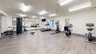 Located at 200 Valencia Dr SE in Albuquerque, New Mexico, this fitness center offers a well-appointed, single-level workout space designed to support a comprehensive exercise routine. The room features durable wood-look flooring throughout, providing a clean and practical surface suited for a wide range of physical activities.

The space is equipped with an assortment of cardio machines, including a treadmill, elliptical trainer, stationary bikes, and a rowing machine. A multi-function cable and weight machine, along with a weight bench and free weights, rounds out the strength training options. Informational fitness posters are mounted along the walls, offering exercise guidance and anatomical references.

Large wall-mounted mirrors span one side of the room, enhancing the sense of openness and allowing users to monitor their form during workouts. Bright, recessed fluorescent lighting ensures excellent visibility throughout the space. A wall-mounted mini-split HVAC unit provides climate control for year-round comfort.

A floor-to-ceiling glass partition with sleek black framing separates the main fitness area from an adjacent room, which appears to function as an office or wellness support space, adding a professional, modern aesthetic to the overall environment. A wall-mounted television is also present for entertainment during workouts. The layout is open and uncluttered, maximizing usable workout floor space.
