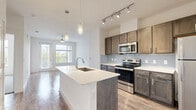 Located at 2700 Babcock Rd in San Antonio, TX, this one-bedroom, one-bathroom unit offers a thoughtfully designed single-floor layout that maximizes every square foot. The open-concept living and kitchen area features warm hardwood-style flooring throughout, creating a cohesive and inviting atmosphere. The kitchen is a standout, equipped with dark walnut-toned shaker cabinets, a subway tile backsplash, light quartz countertops, and a full suite of stainless steel appliances including a refrigerator, electric range, and microwave. A central island with a built-in sink and pendant lighting above provides additional workspace and casual dining potential. Track lighting further brightens the kitchen area. The bedroom is a calm retreat with plush carpeting, a ceiling fan with light fixture, and a window that frames views of surrounding greenery, filling the room with natural light. The bathroom is well-appointed with wood-toned cabinetry, granite countertops, hardwood-style flooring, and a glass-enclosed shower. A dedicated closet with a built-in hanging rod connects directly off the bathroom, offering convenient storage. Clean white walls and bright finishes throughout give the unit a fresh, modern feel.