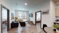 Located at 1 Chevy Chase Lane in Eugene, Oregon, this single-floor unit offers a thoughtfully designed open-concept layout that seamlessly connects the kitchen, dining, and living areas. The space features wood-style flooring throughout, recessed lighting, and crisp white walls that create a clean, contemporary aesthetic.

The kitchen is well-appointed with light wood upper cabinetry, dark lower cabinetry, white countertops, a subway tile backsplash, and a full suite of stainless steel appliances including a refrigerator, range, microwave, and dishwasher. A compact dining area with a round table sits adjacent to the kitchen, making it ideal for everyday meals.

The living area is generously sized, featuring large windows with horizontal blinds that allow ample natural light and views of the surrounding neighborhood. The space comfortably accommodates a sofa, accent chair, coffee table, and wall-mounted television.

The bedroom is bright and serene, with wide windows, wood-style flooring, and room for a full-sized bed and a dedicated desk area — a practical detail well-suited for studying or working from home.

Both bathrooms maintain the modern design language of the unit, featuring dark vanity cabinetry, white countertops, large mirrors, and tub-shower combinations with clean finishes. The overall ambiance is modern, functional, and well-suited for everyday living in Eugene.