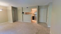 This single-floor space offers a practical and open layout designed for comfortable everyday living. The main living area features neutral-toned carpeting that extends across a generously sized floor plan, creating a warm and inviting atmosphere. Soft sage-green walls contribute to a calm, understated aesthetic throughout.

The kitchen is positioned centrally and is accessible via a tiled entryway, with a half-wall pass-through counter connecting it to the living space — a functional design that promotes an open feel while defining each area. Inside the kitchen, warm-toned wooden cabinetry lines the upper and lower walls, providing ample storage. Stainless steel appliances, including a refrigerator and range, offer modern utility, while tile flooring keeps the cooking area easy to maintain.

A white paneled entry door is situated adjacent to the kitchen, providing the main point of access to the unit. Additional doors visible from the living area suggest access to further rooms or storage. Overhead HVAC venting is integrated into the ceiling, ensuring climate comfort. The overall ambiance is clean, functional, and move-in ready.