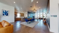 This single-floor community clubhouse showcases a thoughtfully designed open-concept layout that seamlessly connects multiple functional zones. The striking herringbone-patterned flooring flows consistently throughout the entire space, anchoring each area with a warm, cohesive aesthetic.

The welcoming entryway features a dark accent wall, decorative globe pendant lighting, and glass doors that flood the space with natural light. This transitions into a spacious lounge area furnished with tan leather sofas, complemented by a dramatic stacked stone fireplace feature wall, pendant lighting, and tall windows that frame exterior views.

The fully equipped kitchen is outfitted with rich warm-toned wood cabinetry, white subway tile backsplash, quartz countertops, stainless steel appliances, and a large island with bar stool seating — ideal for communal gatherings and events.

The central dining and gathering area includes a wood dining table with bold blue upholstered chairs beneath an elegant orb chandelier. Adjacent to this, a vibrant blue accent wall defines the game room zone, complete with a foosball table and casual seating.

A separate lounge nook, highlighted by floor-to-ceiling windows overlooking an outdoor pool area, offers a bright and relaxing retreat with a sectional sofa. Throughout the space, recessed lighting, curated artwork, and indoor greenery contribute to a polished and inviting communal atmosphere.