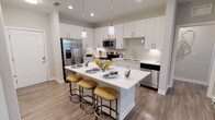 Located at 12851 Sunstone Ave in Orlando's Lake Nona area, this single-floor residence showcases a thoughtfully designed open-concept layout with modern finishes throughout. The kitchen serves as a central highlight, featuring white shaker cabinetry extending to the ceiling, a large quartz-topped island with seating for multiple guests, stainless steel appliances, a subway tile backsplash, and pendant lighting. A dedicated pantry closet and wood-look flooring add both function and style to the space.

The living area is defined by warm golden-yellow accent walls, large windows that invite abundant natural light, and direct access to an outdoor area via a glass-panel door. The open floor plan creates a seamless flow between the kitchen and living spaces.

The primary bedroom offers outdoor views through generous windows, carpeted flooring, and a walk-in closet, while the en-suite bathroom features dual sinks, a walk-in glass shower, and wood-look tile flooring. The secondary bedroom maintains a similarly comfortable atmosphere, complemented by a full bathroom with a tub-shower combination and white vanity cabinetry. Throughout, the design balances contemporary aesthetics with practical functionality.