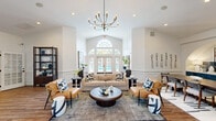 Located at 1201 Aragon Dr in Knightdale, NC, this generously sized single-floor clubhouse offers a well-rounded selection of amenities across a substantial footprint. The centerpiece of the interior is a grand lounge and living area distinguished by soaring vaulted ceilings, a striking multi-arm chandelier, and elegant wainscoting along crisp white walls. A large arched window above French doors floods the space with natural light and frames views of the outdoor pool area, creating a bright and welcoming atmosphere. Rich hardwood flooring anchors the room, which is styled with contemporary furnishings and thoughtful décor. The kitchen features bold teal cabinetry, stainless steel appliances, and warm wood-tone flooring, seamlessly connecting to a dining area framed by a charming arched passageway. The dedicated office space is painted in deep navy blue, accented by crown molding, a glass-paned door, and natural window light — providing a focused, professional environment. The fitness center is well-equipped with strength training machines, free weights, rubberized flooring, and wall-to-wall mirrors. Entry areas feature tile flooring, package lockers, and secure access doors. Outdoors, a large resort-style pool is surrounded by a spacious sundeck, pergola, and lounge seating.