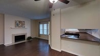 This single-story Dallas space offers an open layout that seamlessly connects the living area and kitchen. The living room features rich dark hardwood flooring, a tiled fireplace with a classic white mantle, and a ceiling fan that provides both function and casual style. Neutral beige walls create a clean, versatile backdrop throughout. A window dressed with horizontal blinds and curtain panels allows natural light to filter into the living area, brightening the space. A half-wall partition with dark countertop separates the kitchen from the living room, creating a defined yet open pass-through arrangement. The kitchen includes a stainless steel sink and a gas or electric stovetop, with an additional window above the sink providing supplemental natural light. The textured popcorn ceiling adds a traditional touch characteristic of the era of construction. Crown molding along the baseboards provides a subtle finishing detail. The overall ambiance is functional and straightforward, with an easy-flowing single-floor layout that suits everyday living in Dallas.