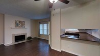This single-story Dallas space offers an open layout that seamlessly connects the living area and kitchen. The living room features rich dark hardwood flooring, a tiled fireplace with a classic white mantle, and a ceiling fan that provides both function and casual style. Neutral beige walls create a clean, versatile backdrop throughout. A window dressed with horizontal blinds and curtain panels allows natural light to filter into the living area, brightening the space. A half-wall partition with dark countertop separates the kitchen from the living room, creating a defined yet open pass-through arrangement. The kitchen includes a stainless steel sink and a gas or electric stovetop, with an additional window above the sink providing supplemental natural light. The textured popcorn ceiling adds a traditional touch characteristic of the era of construction. Crown molding along the baseboards provides a subtle finishing detail. The overall ambiance is functional and straightforward, with an easy-flowing single-floor layout that suits everyday living in Dallas.