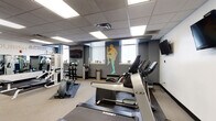 Located at 226 Eighth St E in Cincinnati, OH, this fitness center is a comprehensively outfitted single-floor facility designed to support a full range of workout routines. The space is organized into distinct zones, each serving a specific training purpose. The cardio area features multiple treadmills by Landice, elliptical cross-trainers, and stationary bikes, all positioned beneath a drop-panel ceiling with bright recessed lighting. Multiple wall-mounted flat-screen TVs are positioned throughout for entertainment during workouts. A dedicated free weight area is clearly labeled with bold, colorful wall graphics and includes a full rack of dumbbells and an adjustable weight bench, providing ample options for strength training. An adjacent section houses resistance bands, yoga mats, and a balance scale, catering to flexibility and wellness routines. Teal accent walls and vibrant silhouette graphics of athletes add an energetic and motivating ambiance throughout the space. Natural light filters in through windows fitted with vertical blinds on one side of the room, while rubber flooring mats protect the workout areas. French doors with glass panes provide an elegant entry point into the facility, contributing a refined architectural touch to an otherwise functional, performance-focused environment.