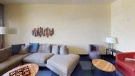 This single-story clubhouse is designed to offer residents a comfortable and functional communal gathering space. The interior is characterized by warm cream-colored walls accented by bold blue feature walls, creating a visually engaging environment throughout. Blue-toned checkerboard-patterned carpet tiles unify the space while adding a modern design element underfoot.

The lounge area serves as the social hub of the clubhouse, furnished with a generously sized sectional sofa in neutral gray, complemented by blue accent pillows and a striking mixed-media wall sculpture featuring wood and red accents. Additional seating includes ribbed barrel chairs and round wood-topped side tables, creating a relaxed yet contemporary atmosphere. An oversized arc floor lamp provides ambient lighting alongside natural light from windows.

A dedicated dining nook features a round dark wood table surrounded by upholstered chairs, positioned directly beside a large floor-to-ceiling window that frames outdoor greenery and landscaping, flooding the area with natural light.

The entry area includes a welcoming seating vignette with a red accent chair, a side table with a table lamp, and glass-panel doors leading further into the facility, providing a polished and inviting first impression.