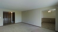 This single-floor space presents a straightforward, functional layout with a notably open living area. The main room features neutral beige walls paired with wall-to-wall carpeting in a matching light tone, creating a cohesive and understated aesthetic. Dark baseboards provide a subtle contrast against the lighter walls and flooring. The ceiling is smooth and white, fitted with a central HVAC vent, contributing to a clean, uncluttered overhead appearance. A doorway on the left side of the main room leads to an adjacent space, likely a closet or secondary room, while an open passage on the right connects to what appears to be a dining or transitional area. This adjoining space features a distinctive chair-rail detail along the walls, adding a modest architectural element. The layout suggests a practical flow between living and dining zones. Overall, the space presents a blank canvas with a calm, neutral palette, making it highly adaptable for various furnishing and décor styles. The absence of windows visible in the image suggests a more enclosed interior orientation.