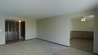 This single-floor space presents a straightforward, functional layout with a notably open living area. The main room features neutral beige walls paired with wall-to-wall carpeting in a matching light tone, creating a cohesive and understated aesthetic. Dark baseboards provide a subtle contrast against the lighter walls and flooring. The ceiling is smooth and white, fitted with a central HVAC vent, contributing to a clean, uncluttered overhead appearance. A doorway on the left side of the main room leads to an adjacent space, likely a closet or secondary room, while an open passage on the right connects to what appears to be a dining or transitional area. This adjoining space features a distinctive chair-rail detail along the walls, adding a modest architectural element. The layout suggests a practical flow between living and dining zones. Overall, the space presents a blank canvas with a calm, neutral palette, making it highly adaptable for various furnishing and décor styles. The absence of windows visible in the image suggests a more enclosed interior orientation.