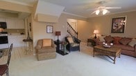 This two-story home centers around a spacious living area finished with plush wall-to-wall carpeting and warm neutral-toned walls that create an inviting atmosphere throughout. Crown molding adds a refined architectural touch, complementing the angled ceiling detail visible above. A ceiling fan with a light fixture provides both comfort and ambient illumination. The living area offers generous room for multiple seating arrangements, with the layout naturally guiding movement toward the staircase, which features a classic dark wood banister and spindle railing leading to the upper level. The staircase serves as a visual focal point within the open layout, elegantly connecting the two floors. The warm earth-toned palette is carried consistently throughout the space, lending a cohesive and comfortable feel. Accent lighting from table lamps adds a welcoming glow, enhancing the overall ambiance. The combination of architectural detailing, multi-level design, and well-proportioned living spaces makes this home both functional and aesthetically appealing.