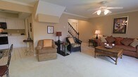 This two-story home centers around a spacious living area finished with plush wall-to-wall carpeting and warm neutral-toned walls that create an inviting atmosphere throughout. Crown molding adds a refined architectural touch, complementing the angled ceiling detail visible above. A ceiling fan with a light fixture provides both comfort and ambient illumination. The living area offers generous room for multiple seating arrangements, with the layout naturally guiding movement toward the staircase, which features a classic dark wood banister and spindle railing leading to the upper level. The staircase serves as a visual focal point within the open layout, elegantly connecting the two floors. The warm earth-toned palette is carried consistently throughout the space, lending a cohesive and comfortable feel. Accent lighting from table lamps adds a welcoming glow, enhancing the overall ambiance. The combination of architectural detailing, multi-level design, and well-proportioned living spaces makes this home both functional and aesthetically appealing.