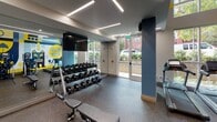 Located on Poindexter Drive in Charlotte's dynamic Southend neighborhood, this two-floor fitness center combines functional design with energetic, locally inspired aesthetics. The space is immediately distinguished by a bold, large-scale mural rendered in navy blue, yellow, and gray — depicting iconic Southend imagery including cyclists, roller skaters, and neighborhood landmarks — that wraps along the walls and sets a lively, community-rooted tone throughout.

The fitness floor is equipped with a comprehensive array of Matrix-brand machines, including ellipticals, treadmills, rowing machines, and a multi-use cable and weight system. A full rack of dumbbells and a dedicated free-weight bench offer versatile strength training options, while exercise balls provide space for functional fitness work. Floor-to-ceiling mirrors line key sections of the space, visually expanding the area and supporting form-focused training.

Linear LED lighting runs across the ceiling in clean, modern strips, ensuring the space is well-lit throughout. Large glass doors and windows allow natural light to pour in, offering views of the surrounding streetscape and greenery. The flooring consists of durable, speckled rubber material suited for high-traffic athletic use.

A spiral staircase connects the two floors, and the layout also includes additional rooms and utility areas accessible via interior doors. The overall ambiance is energetic yet polished, making this fitness center a standout amenity in the Southend community.