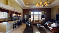 Clubhouse East at 45418 Howe Rd in San Diego is a single-floor community gathering space designed with a sophisticated and welcoming aesthetic. The entrance greets visitors with soaring ceilings, warm neutral-toned walls, and large arched glass doors that draw in natural light and create an open, airy atmosphere. Light tile flooring flows seamlessly throughout the space, unifying each distinct area.

The main lounge is a standout feature, boasting dramatically tall walls painted in deep chocolate brown, floor-to-ceiling windows with layered curtains and wood blinds, and generous natural light. Comfortable seating arrangements — including plush sofas and leather chairs — are centered around a sleek coffee table, making it ideal for socializing or relaxing.

Adjacent to the lounge is a full kitchen, outfitted with rich dark wood cabinetry, granite countertops, a granite tile backsplash, and stainless steel appliances including a gas range. A breakfast bar with bar seating provides a casual dining option and connects the kitchen to the main living area.

A separate dining nook offers a more intimate setting, furnished with a round dark wood table and chairs beneath a decorative chandelier, with a door leading directly to the outdoors and views of the surrounding landscape. Throughout, crown molding, tray ceilings, and thoughtful lighting fixtures add refined architectural detail to this well-appointed clubhouse.