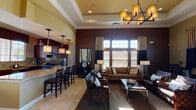 Clubhouse East at 45418 Howe Rd in San Diego is a single-floor community gathering space designed with a sophisticated and welcoming aesthetic. The entrance greets visitors with soaring ceilings, warm neutral-toned walls, and large arched glass doors that draw in natural light and create an open, airy atmosphere. Light tile flooring flows seamlessly throughout the space, unifying each distinct area.

The main lounge is a standout feature, boasting dramatically tall walls painted in deep chocolate brown, floor-to-ceiling windows with layered curtains and wood blinds, and generous natural light. Comfortable seating arrangements — including plush sofas and leather chairs — are centered around a sleek coffee table, making it ideal for socializing or relaxing.

Adjacent to the lounge is a full kitchen, outfitted with rich dark wood cabinetry, granite countertops, a granite tile backsplash, and stainless steel appliances including a gas range. A breakfast bar with bar seating provides a casual dining option and connects the kitchen to the main living area.

A separate dining nook offers a more intimate setting, furnished with a round dark wood table and chairs beneath a decorative chandelier, with a door leading directly to the outdoors and views of the surrounding landscape. Throughout, crown molding, tray ceilings, and thoughtful lighting fixtures add refined architectural detail to this well-appointed clubhouse.