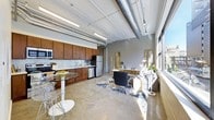 This single-floor space showcases a distinct industrial-loft aesthetic, combining raw architectural details with functional living areas in an open-plan layout. Exposed ductwork and structural beams run across the ceiling, complemented by polished concrete floors that extend throughout the space, lending an urban, warehouse-inspired character.

The kitchen is well-appointed with rich, dark wood cabinetry spanning the full length of the wall, paired with a mosaic tile backsplash and stainless steel appliances, including a refrigerator, dishwasher, and range. A compact bistro-style dining table with modern seating sits adjacent to the kitchen, creating an intimate dining nook.

The living and workspace area benefits from a generous row of large windows along one wall, flooding the interior with natural light and offering views of the surrounding urban environment. A dedicated workspace features a desk and seating arrangement, while decorative elements including a patterned area rug, indoor plants, and curated accessories add warmth to the otherwise industrial palette. Linear fluorescent pendant lighting provides additional illumination throughout.