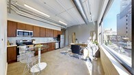 This single-floor space showcases a distinct industrial-loft aesthetic, combining raw architectural details with functional living areas in an open-plan layout. Exposed ductwork and structural beams run across the ceiling, complemented by polished concrete floors that extend throughout the space, lending an urban, warehouse-inspired character.

The kitchen is well-appointed with rich, dark wood cabinetry spanning the full length of the wall, paired with a mosaic tile backsplash and stainless steel appliances, including a refrigerator, dishwasher, and range. A compact bistro-style dining table with modern seating sits adjacent to the kitchen, creating an intimate dining nook.

The living and workspace area benefits from a generous row of large windows along one wall, flooding the interior with natural light and offering views of the surrounding urban environment. A dedicated workspace features a desk and seating arrangement, while decorative elements including a patterned area rug, indoor plants, and curated accessories add warmth to the otherwise industrial palette. Linear fluorescent pendant lighting provides additional illumination throughout.