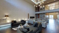 This sophisticated modern lounge makes an immediate impression with its soaring double-height ceilings and an expansive, floor-to-ceiling textured white accent wall that serves as the room's dramatic focal point. Subtle uplighting accentuates the wall's undulating surface, adding depth and warmth to the space. Anchoring the wall is a sleek, linear gas fireplace framed by an ornate vertical tile inset, blending contemporary and eclectic design sensibilities seamlessly.

Overhead, a sculptural chandelier composed of clustered globe pendants on branching brass arms casts a warm, ambient glow throughout the room. Dark hardwood flooring grounds the space, contrasting elegantly with the light tones of the walls and furnishings. Multiple seating arrangements — featuring a mix of charcoal sofas, light gray sectionals, and upholstered accent chairs — provide flexible, comfortable gathering areas suited for both intimate conversations and larger groups.

An upper-level mezzanine with a modern metal railing is visible above, overlooking the main living area and enhancing the sense of vertical grandeur. Decorative Moorish-inspired mirror panels adorn one wall, adding an artistic, globally influenced touch. The overall ambiance is one of refined luxury — modern in structure, yet layered with curated details that make the space feel inviting and design-forward.