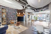 Located at 318 I St NE in Washington, DC, this single-floor amenity space at The Axis offers a thoughtfully designed, open-plan interior that blends contemporary aesthetics with functional versatility. The polished concrete floors reflect abundant natural light streaming through expansive floor-to-ceiling glass walls and doors, which also provide views of an outdoor courtyard area. Exposed ductwork and industrial-style ceilings contrast beautifully with sculptural curved ceiling elements that add architectural interest overhead. A bold, colorful graphic feature wall serves as a striking focal point near the entrance, complemented by built-in shelving displaying curated décor. The space accommodates a variety of uses, with a casual lounge area furnished with rounded blue velvet chairs and bistro-style tables, as well as a more formal conference zone featuring a large oval table surrounded by upholstered seating and a wall-mounted display screen framed by a textured stone accent wall. A reception counter anchors the central area, while pendant lighting and warm wood wall panels add depth and warmth throughout. Lush potted plants are positioned throughout, enhancing the welcoming, vibrant ambiance of this well-appointed community space.