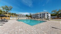 This outdoor amenity space centers around a generously sized rectangular swimming pool with clear blue water, offering an inviting retreat for residents. The pool area is finished with elegant paver decking that surrounds the water on all sides, providing ample space for lounging and socializing. Rows of chaise lounge chairs are thoughtfully arranged along the pool's perimeter, with market umbrellas offering shaded seating options for those seeking relief from the sun.

A standout architectural feature is the wooden pergola structure positioned at one end of the pool, adding a warm, natural aesthetic element to the outdoor environment. An additional pergola accent enhances the social atmosphere of the deck. The pool itself includes clearly marked depth indicators and easy-access entry points with handrails.

The perimeter of the amenity space is defined by a black wrought iron fence, providing both security and an open, airy feel. A support structure — likely a pool house or storage facility — is situated adjacent to the deck, finished in crisp white with a modern barn-style roof. Mature trees line the backdrop, offering natural privacy screening. Curved walkways connect the pool area to the broader residential community, which features multi-story buildings with classic architectural detailing, balconies, and pitched rooflines visible in the background.
