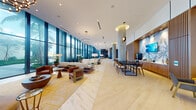 This single-floor lobby is a masterclass in modern design, characterized by its generous proportions and seamless flow between functional zones. Polished marble-style tile flooring runs throughout the main corridor, while select areas transition to light wood herringbone parquet, adding warmth and visual contrast. Soaring floor-to-ceiling glazed walls line one side of the space, flooding the interior with natural light and framing lush tropical landscaping and palm trees beyond.

Substantial concrete columns punctuate the layout, adding architectural character alongside tiered chandelier lighting that provides ambient illumination. A welcoming lounge area features plush sofas, leather armchairs, and a glass coffee table positioned against the expansive glazing. A formal reception desk with woven wood detailing and guest seating anchors a dedicated concierge zone, accented by curated original artwork on adjacent walls.

A dining and meeting area includes a dark conference table with upholstered chairs, while a separate bar-style service station with warm wood cabinetry provides additional functionality. A curved seating nook near the entrance offers an intimate gathering space. Blue-toned accent walls, open shelving, and decorative fixtures complete the refined, resort-inspired atmosphere throughout.
