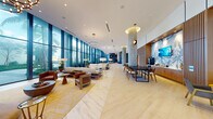 This single-floor lobby is a masterclass in modern design, characterized by its generous proportions and seamless flow between functional zones. Polished marble-style tile flooring runs throughout the main corridor, while select areas transition to light wood herringbone parquet, adding warmth and visual contrast. Soaring floor-to-ceiling glazed walls line one side of the space, flooding the interior with natural light and framing lush tropical landscaping and palm trees beyond.

Substantial concrete columns punctuate the layout, adding architectural character alongside tiered chandelier lighting that provides ambient illumination. A welcoming lounge area features plush sofas, leather armchairs, and a glass coffee table positioned against the expansive glazing. A formal reception desk with woven wood detailing and guest seating anchors a dedicated concierge zone, accented by curated original artwork on adjacent walls.

A dining and meeting area includes a dark conference table with upholstered chairs, while a separate bar-style service station with warm wood cabinetry provides additional functionality. A curved seating nook near the entrance offers an intimate gathering space. Blue-toned accent walls, open shelving, and decorative fixtures complete the refined, resort-inspired atmosphere throughout.